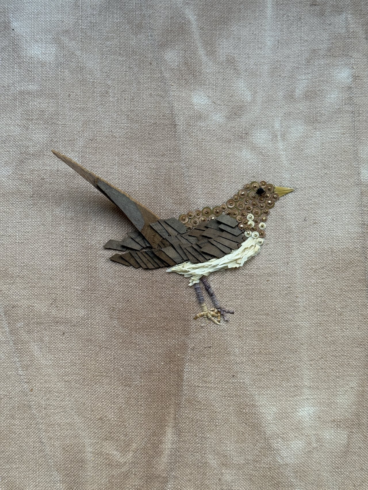 Un oiseau brodé en bois, fil et paillettes sur une toile de lin